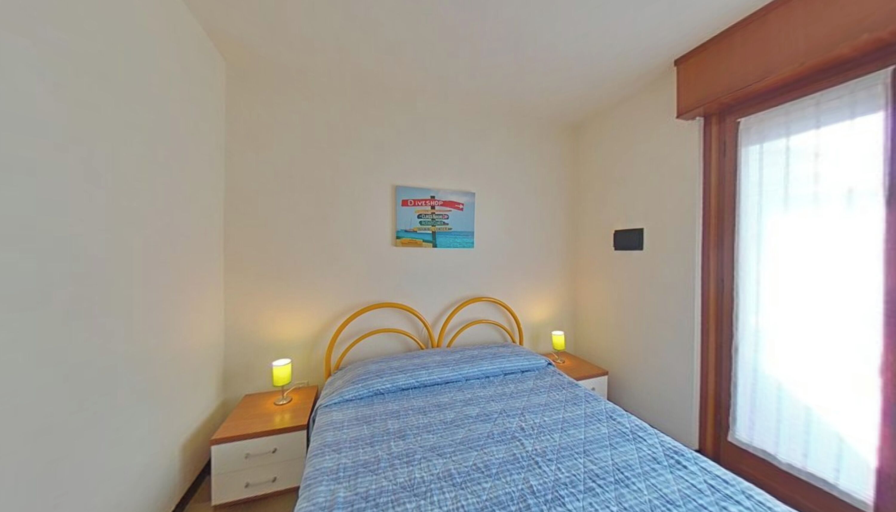 A bedroom with a bed, two bedside tables with lamps, a wall sign, and a window with blinds.