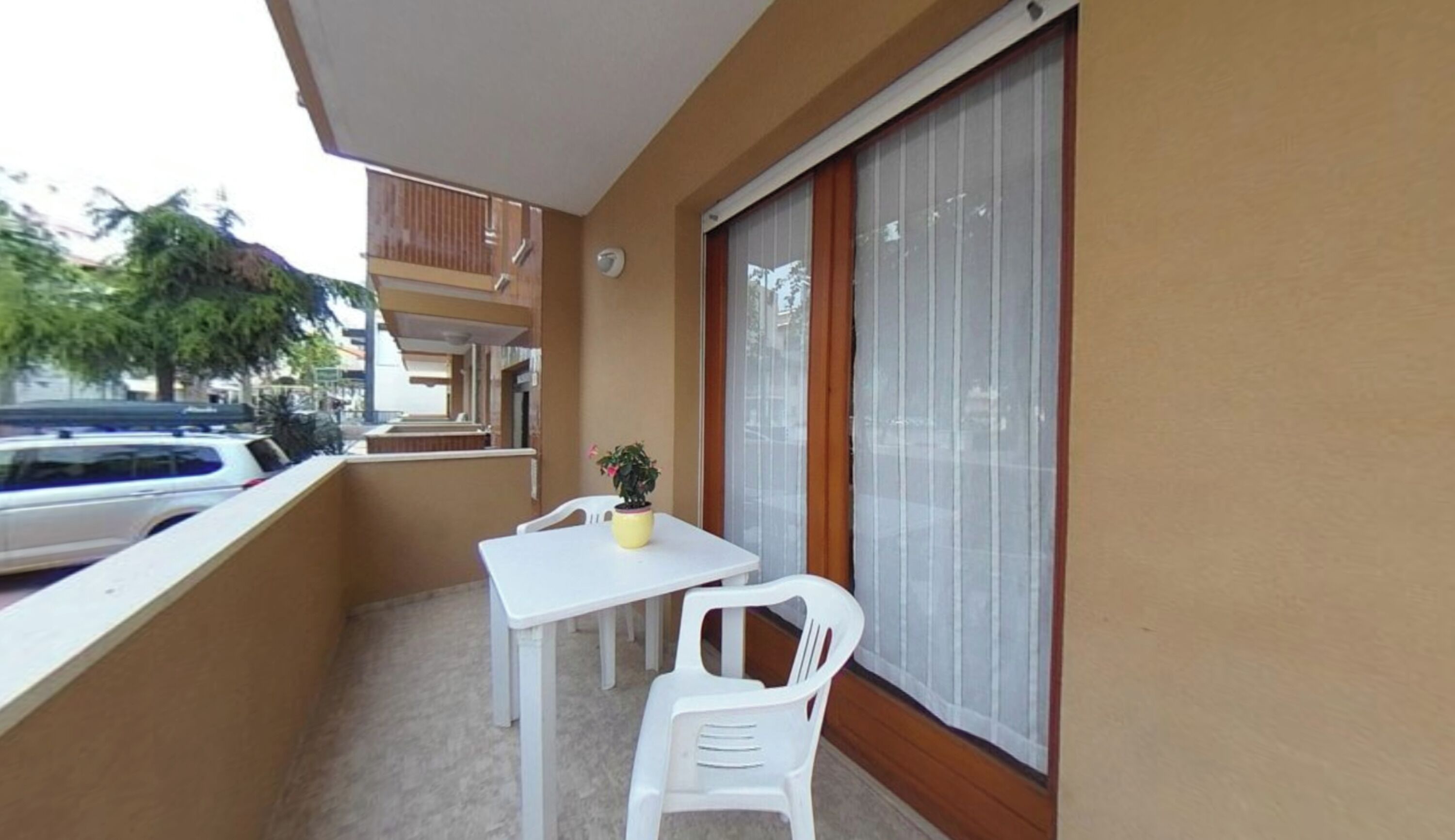 A balcony with a white table and chair, a potted plant, and a view of parked cars.