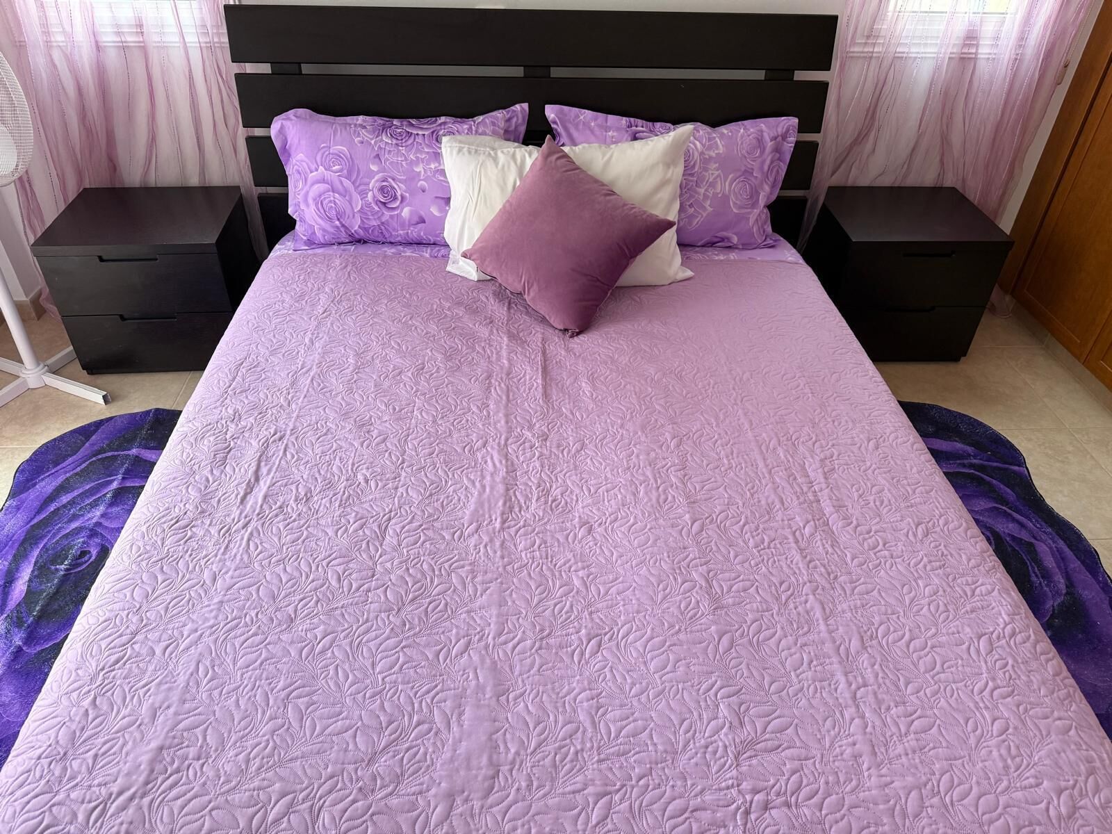 Ein Bett mit einer violetten Steude, zwei Nachttischen und einem Fenster mit durchsichtigen Vorhängen.