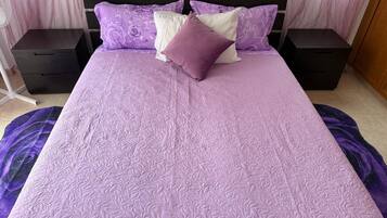 Ein Bett mit einer violetten Steude, zwei Nachttischen und einem Fenster mit durchsichtigen Vorhängen.
