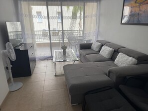 Un salon avec un canapĂ© gris, une table basse en verre, un petit bureau avec un ventilateur, et une vue sur un balcon agrĂ©mentĂ© dâun rideau en dentelle.