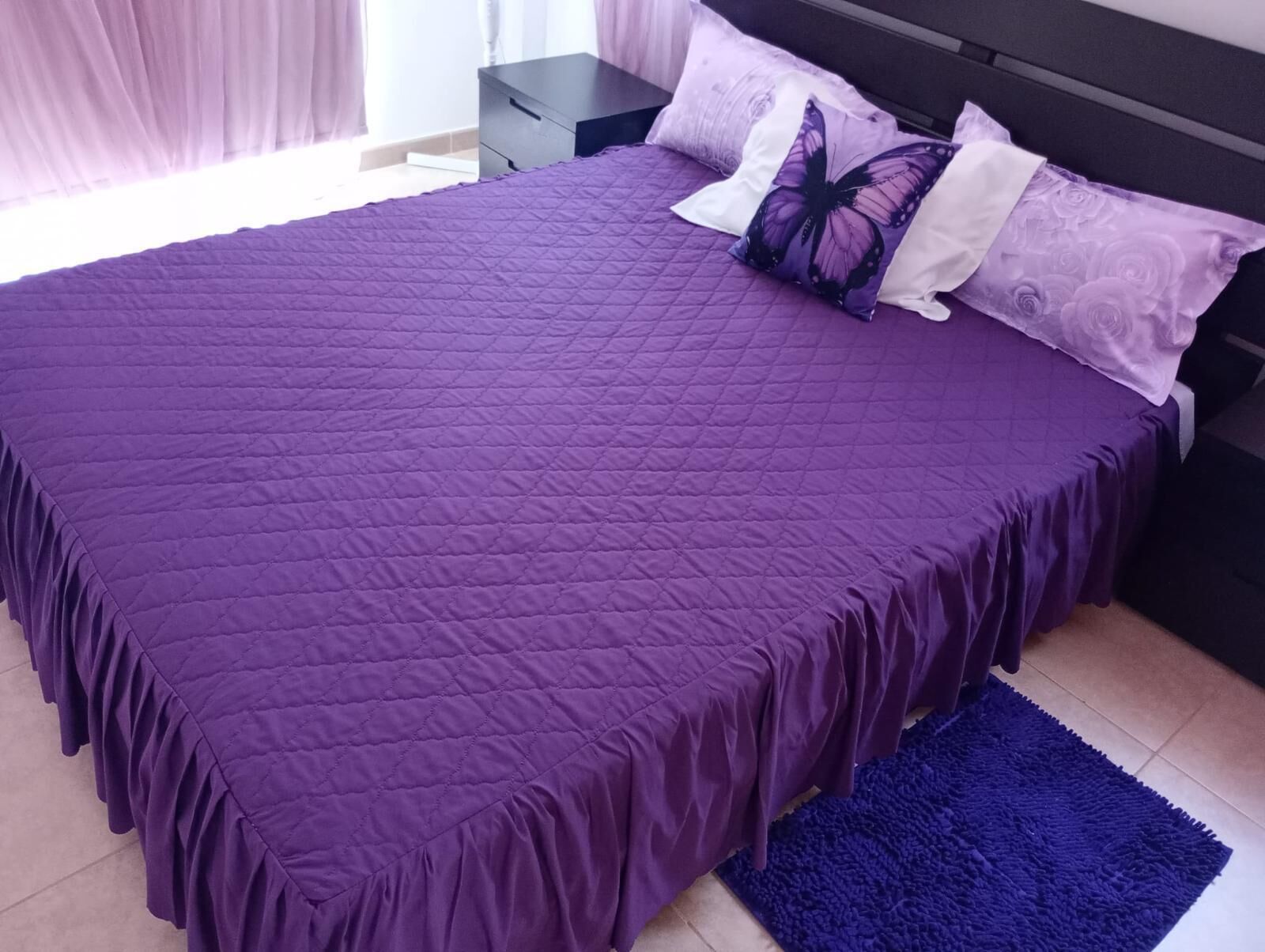 Una cama con un edredón morado, una alfombra azul y una mesita de noche con una lámpara.