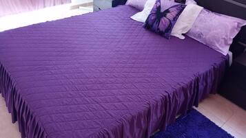 Una cama con un edredón morado, una alfombra azul y una mesita de noche con una lámpara.