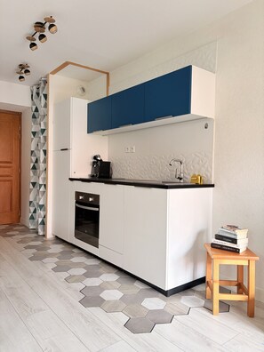 A compact kitchen with white cabinets, a blue upper section, a built-in oven, a sink, and a small wooden stool with books. - Quiet Elegance Near Lake Annecy Giez (Faverges-Seythenex)