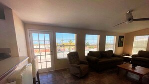 Una sala de estar con sofá, sillón y mesa de centro. Se ve una puerta que da a un patio con vistas al exterior.