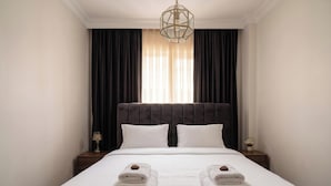 A hotel room with a large bed, dark curtains, a chandelier, and a nightstand with a lamp. - Galata Sea Suites (İstanbul)