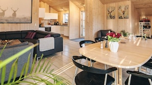 Ein modernes Interieur mit Holzbalkendecke, einem Essbereich mit rundem Tisch und Stühlen, einer Couch und einer Küche im Hintergrund.