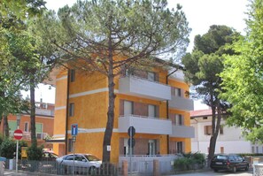 A multi-story building with balconies, a tree in front, and parked cars. - Flat Steps From the sea and Center (Lignano Sabbiadoro)