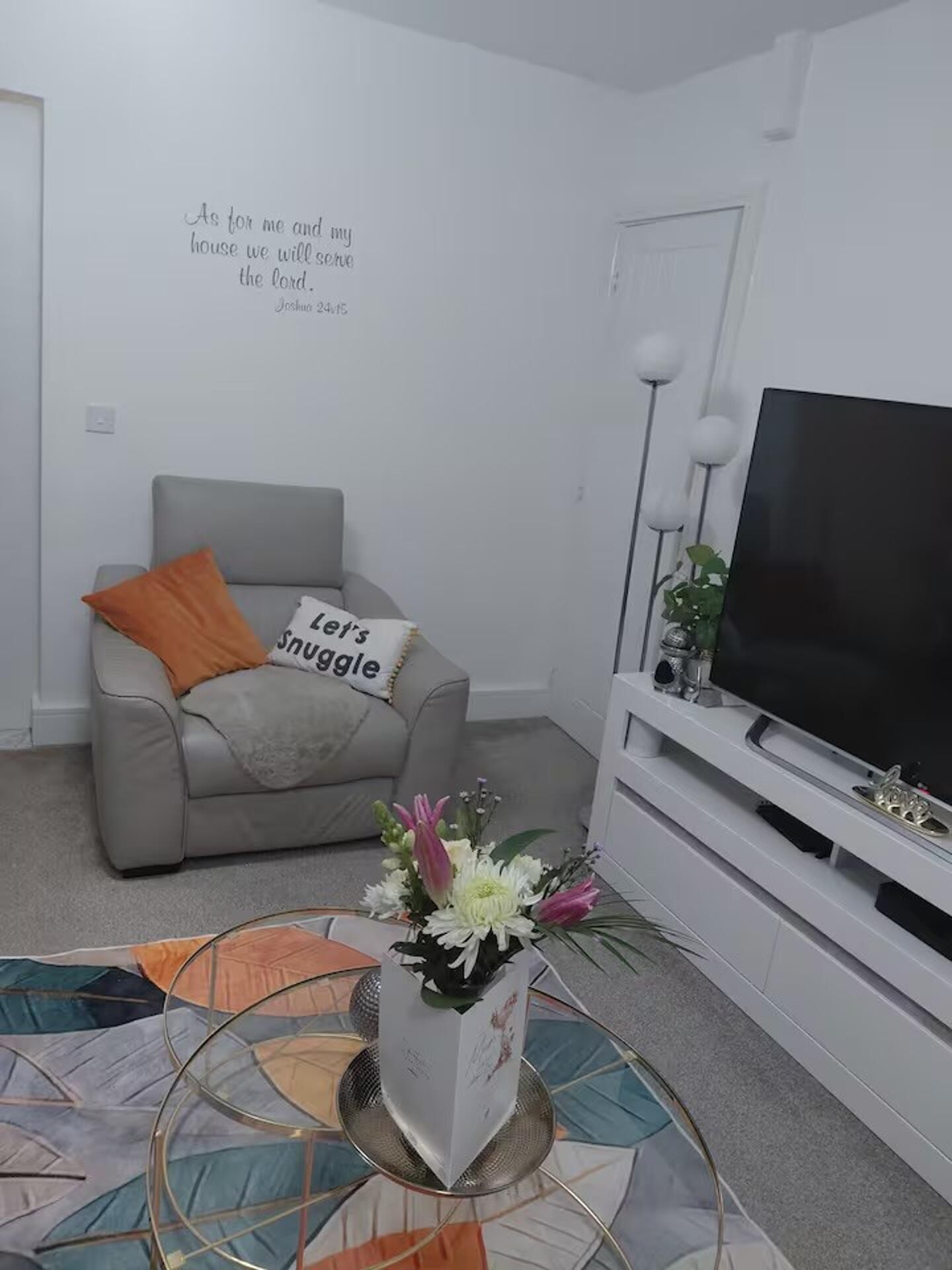 Una sala de estar con un sofá gris, un cojín naranja, un mueble para TV y una cita decorativa en la pared.
