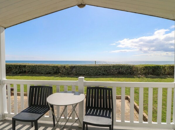 A covered outdoor seating area with a table and two chairs, overlooking a grassy area and the sea. - Caravan 117 (Great Yarmouth)