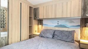 A bedroom with a bed, wooden paneling, a nightstand, a wall-mounted lamp, and a painting of a lighthouse. - Caravan 117 (Great Yarmouth)