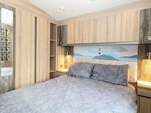 A bedroom with a bed, wooden paneling, a nightstand, a wall-mounted lamp, and a painting of a lighthouse. - Caravan 117 (Great Yarmouth)