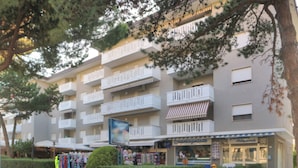 A multi-story building with balconies, a storefront with a sign, and a small garden area. - Practical Flat Near the Beach and Town Center (Lignano Sabbiadoro)
