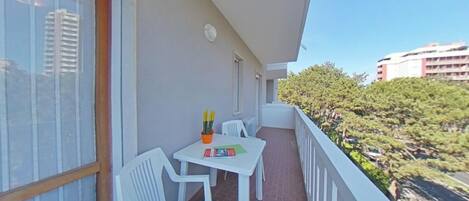 A balcony with a table and chairs, a window with curtains, and a view of buildings and trees.
