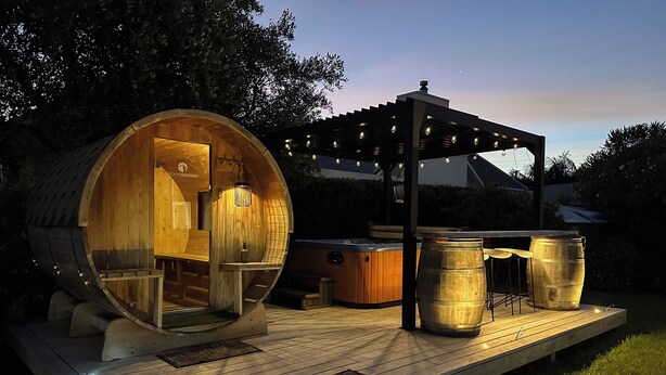 Een houten, vatvormig bouwwerk met een hottub en een pergola met lichtslingers.