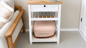 Een klein nachtkastje met een geweven wanddecoratie, een groen hydratatiezakje, een glazen pot en een roze opbergdoos.