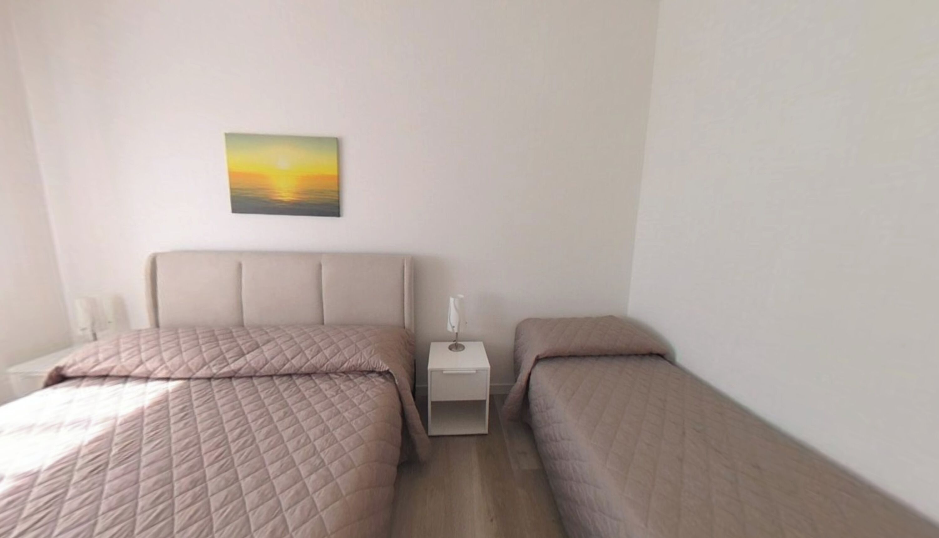 Une chambre avec deux lits, une table de chevet, une lampe fixée au mur et un tableau représentant un coucher de soleil.