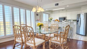 A dining area with a glass table and wicker chairs, a kitchen with white cabinets, and a stainless steel refrigerator. - Cambridge 410 - Beautiful Condo in Litchfield by the Sea (Pawleys Island)