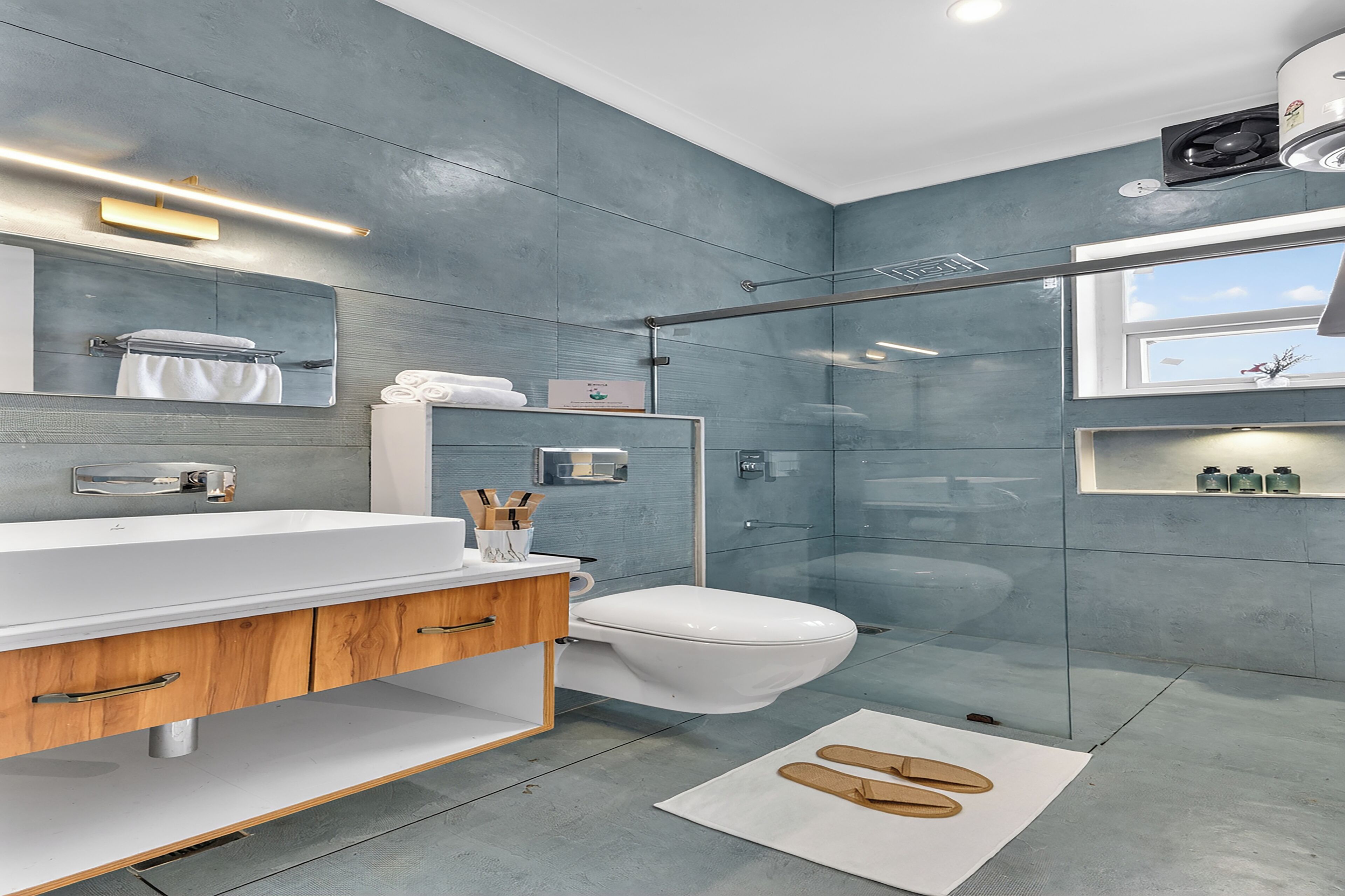 Un baño moderno con un espejo grande, lavamanos blanco, tocador de madera, cabina de ducha de vidrio y vistas al cielo a través de una ventana.