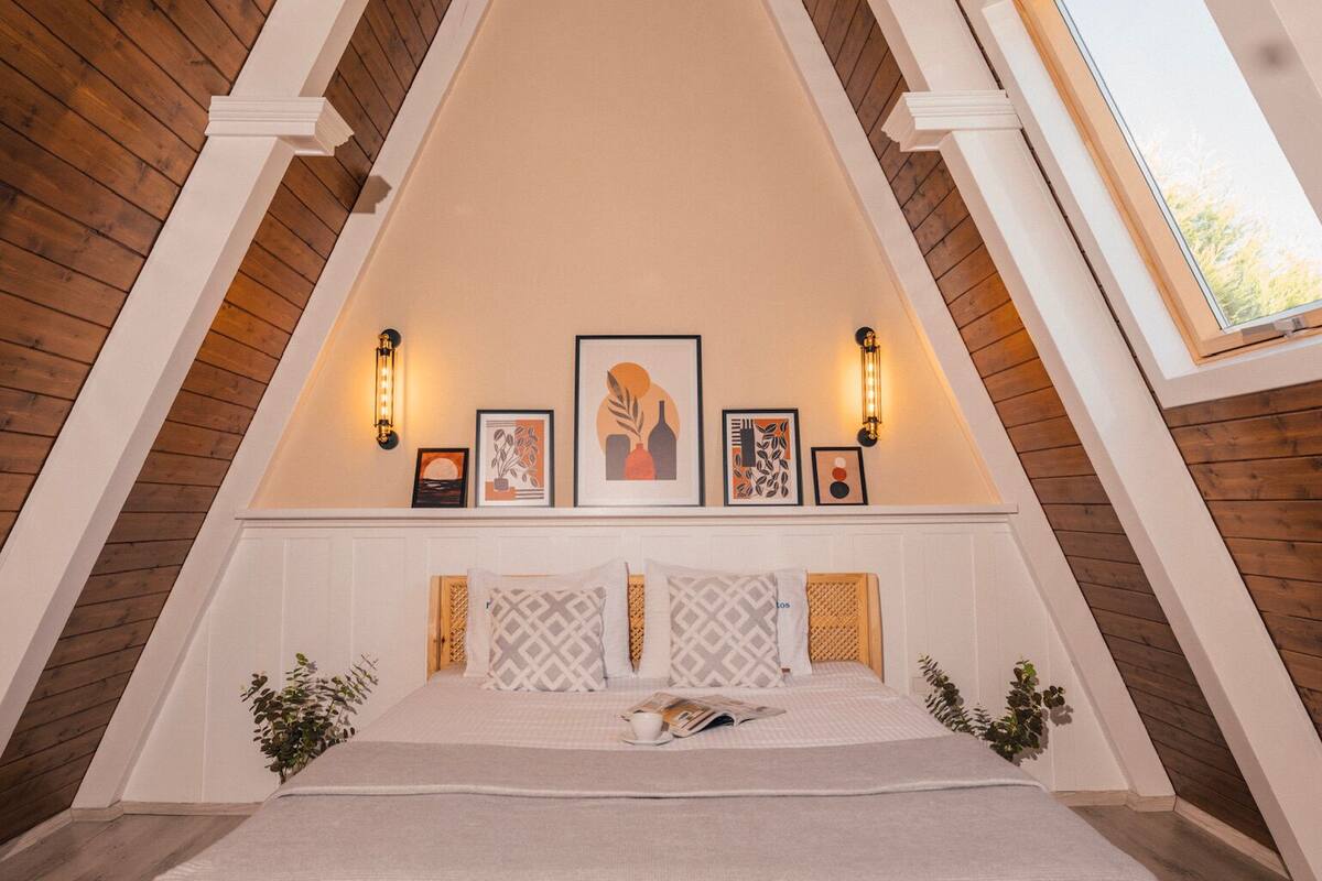 A bedroom with a sloped ceiling, a bed, wall-mounted lights, framed artwork, and a window.