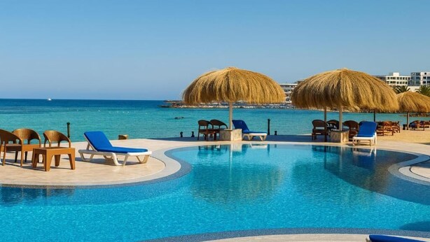 A swimming pool with lounge chairs and thatched umbrellas, surrounded by a clear blue sea and buildings in the background.