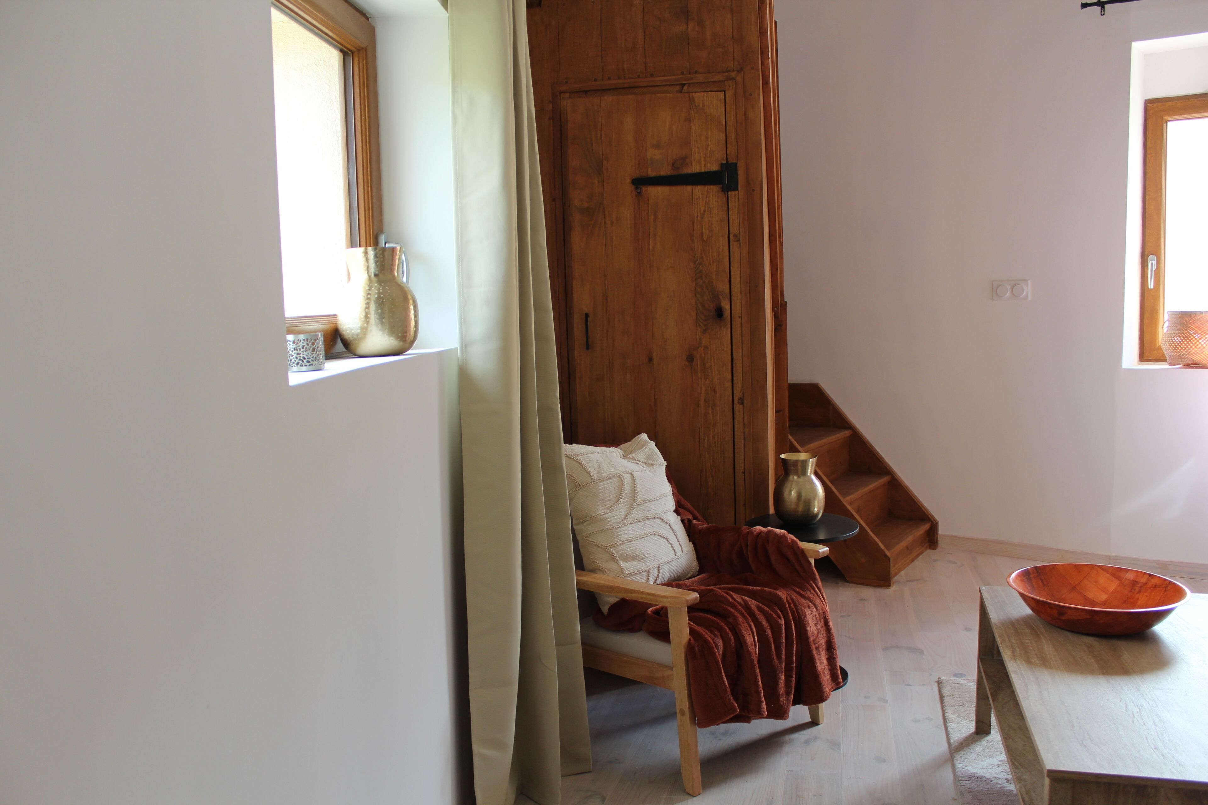 Una habitación con puerta de madera, una silla con manta, una mesa con tazón y una ventana con cortina.