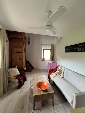 A living room with a wooden floor, a white sofa, a wooden coffee table with a bowl, a ceiling fan, and a wooden door.