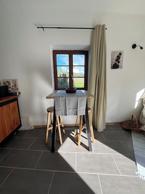 A room with a table, stools, a window with a view, a curtain, a wall-mounted lamp, and a small basket.