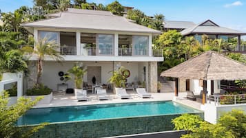 A two-story house with a swimming pool, outdoor seating, and a thatched-roof structure.