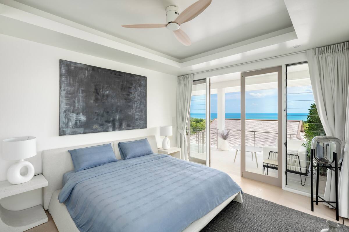 Une chambre à coucher avec un lit, un ventilateur de plafond, une grande fenêtre et une vue sur l’océan.