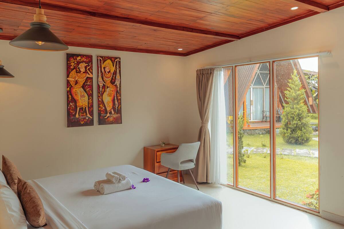 A bedroom with a wooden ceiling, a bed, a desk, and a view of the outdoors.