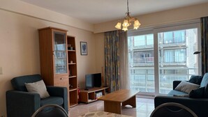 A living room with a sofa, a TV, a cabinet, a table, and a large window with a view of buildings. - Modern Stay With Balcony & Seaview (Middelkerke)