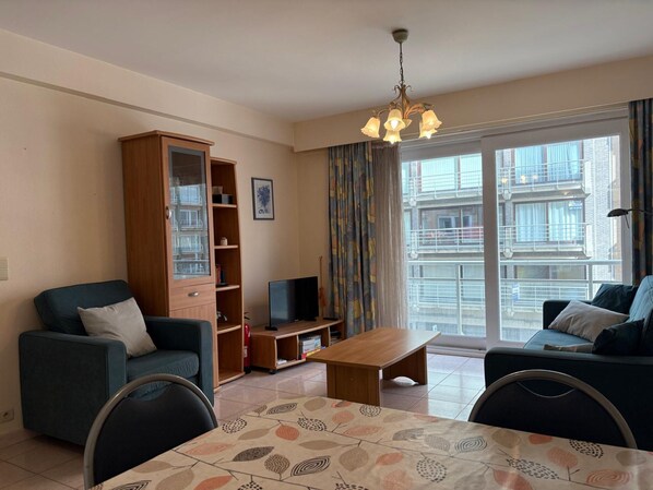 A living room with a sofa, a TV, a cabinet, a table, and a large window with a view of buildings. - Modern Stay With Balcony & Seaview (Middelkerke)