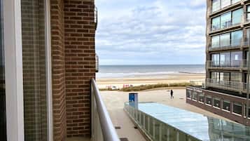 A balcony with a view of the beach, a glass railing, and a building with multiple windows.