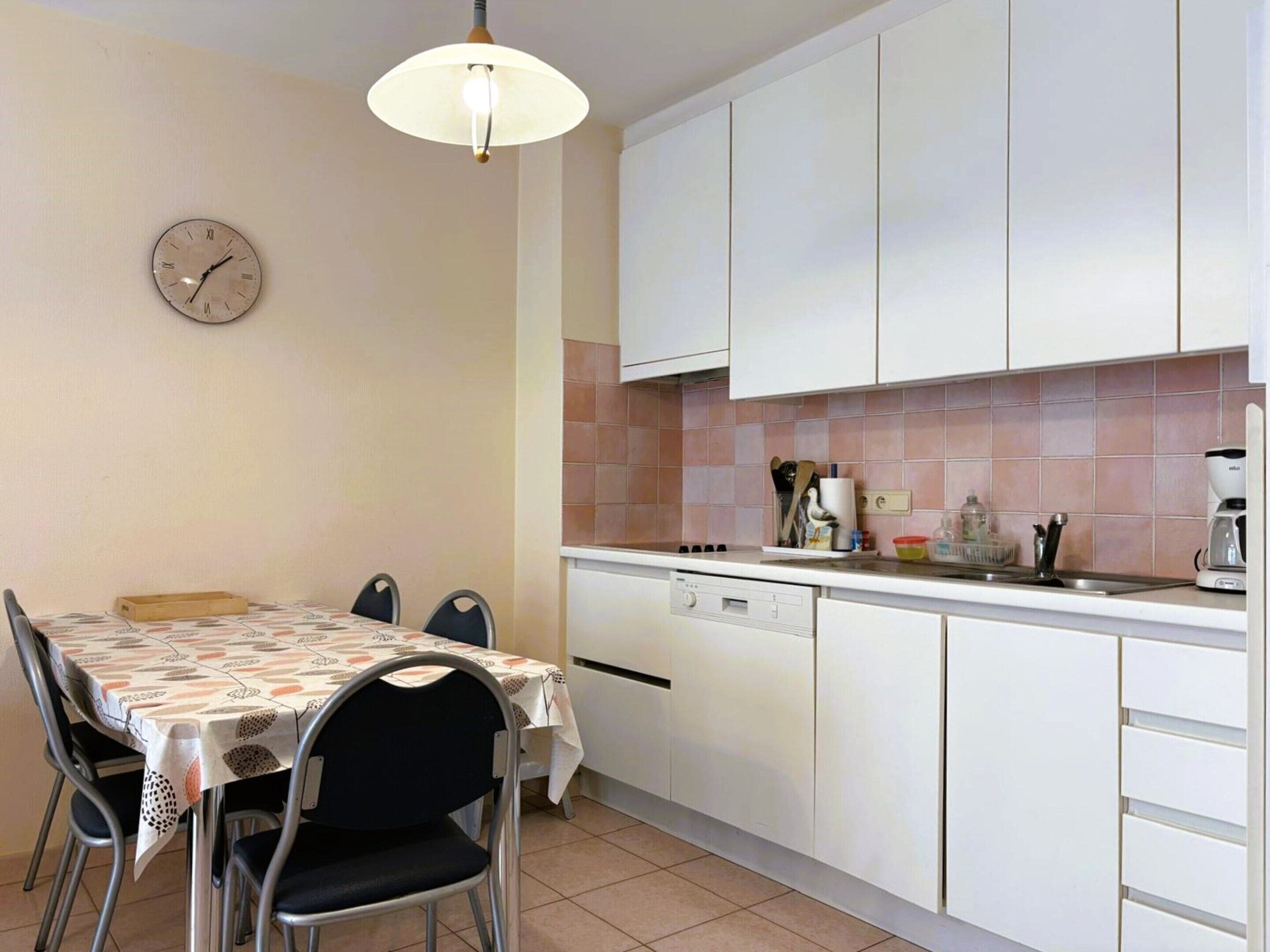A compact kitchen with white cabinets, a dining table with chairs, a clock, and basic kitchen appliances.