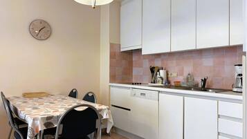 A compact kitchen with white cabinets, a dining table with chairs, a clock, and basic kitchen appliances.