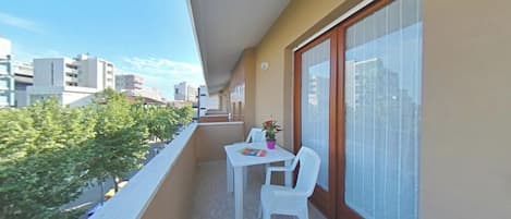 Uma varanda com mesa e cadeiras brancas, um vaso de plantas e vista para a cidade.