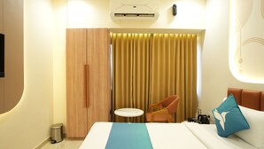A hotel room with a bed, a chair, a small table, an air conditioning unit, and a curtain. - Privilon Hotel Prahladnagar (Ahmedabad)