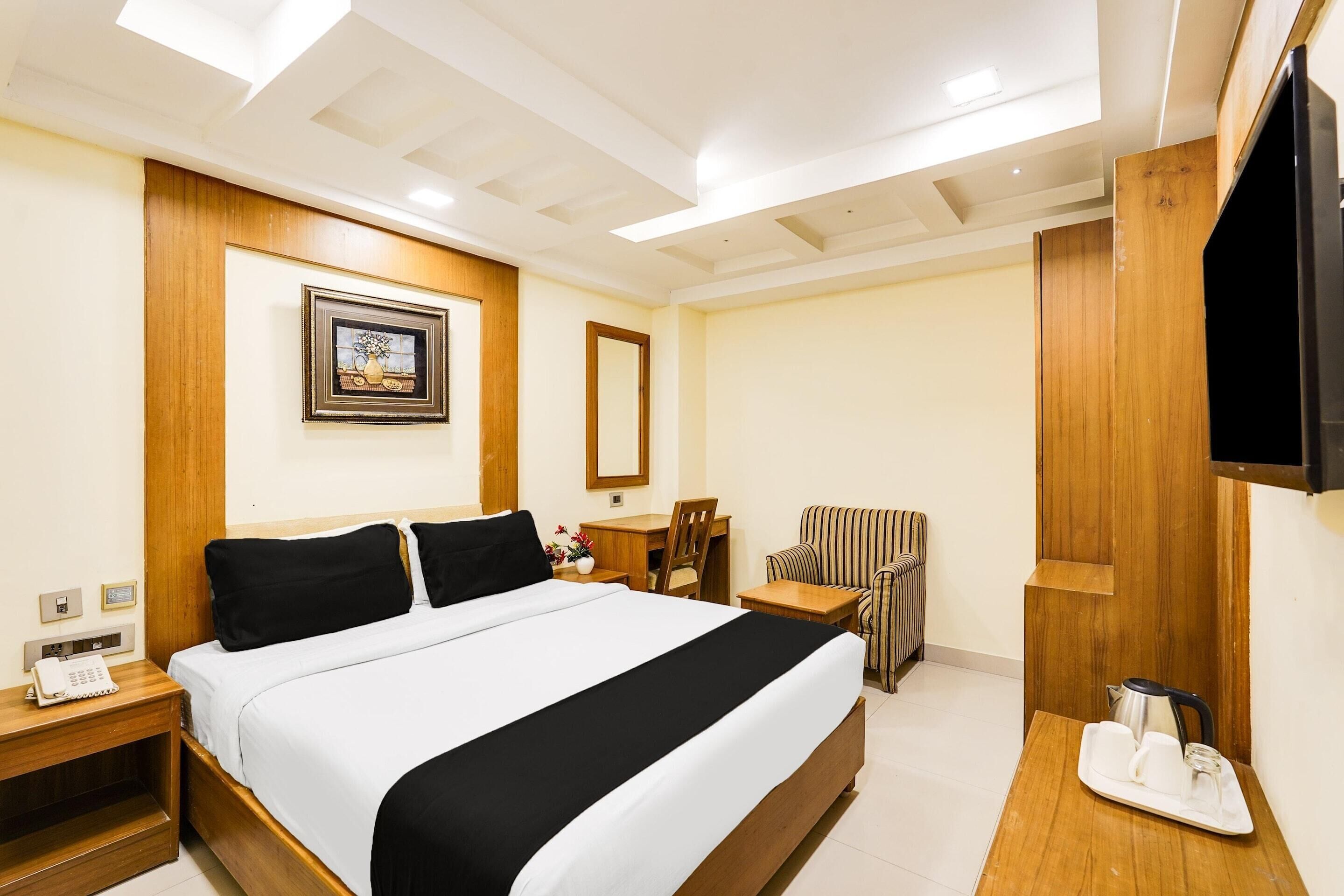 Ein Hotelzimmer mit einem großen Bett, einem Nachttisch aus Holz mit einem Telefon, einem Schreibtisch mit Stuhl, einem an der Wand montierten Fernseher und einem gerahmten Bild.