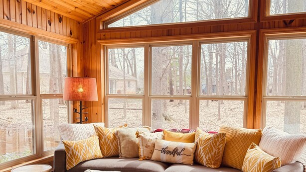 Interior acogedor de una cabaña con techo de madera, ventanas amplias, sofá con cojines estampados y una lámpara.