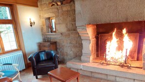A stone fireplace with a fire, a leather sofa, a wooden table, and a window with blinds. - Seaview Stone House in Brittany (Saint-Pol-de-Leon)