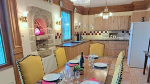 A dining area with a long table set for six, wooden chairs, a stone fireplace, and a kitchen in the background. - Seaview Stone House in Brittany (Saint-Pol-de-Leon)