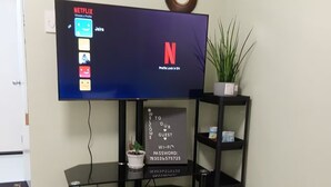 Televisão de tela plana presa a um suporte preto, com interface da Netflix, um sinal de boas-vindas e um vaso com planta.