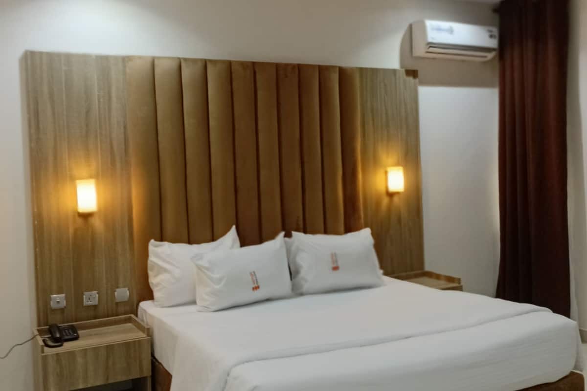 A modern hotel room with a large bed, wooden headboard, bedside lamps, an air conditioning unit, and a nightstand with a phone.