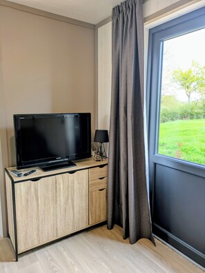A room with a TV on a wooden cabinet, a remote control, a lamp, and a window with curtains.