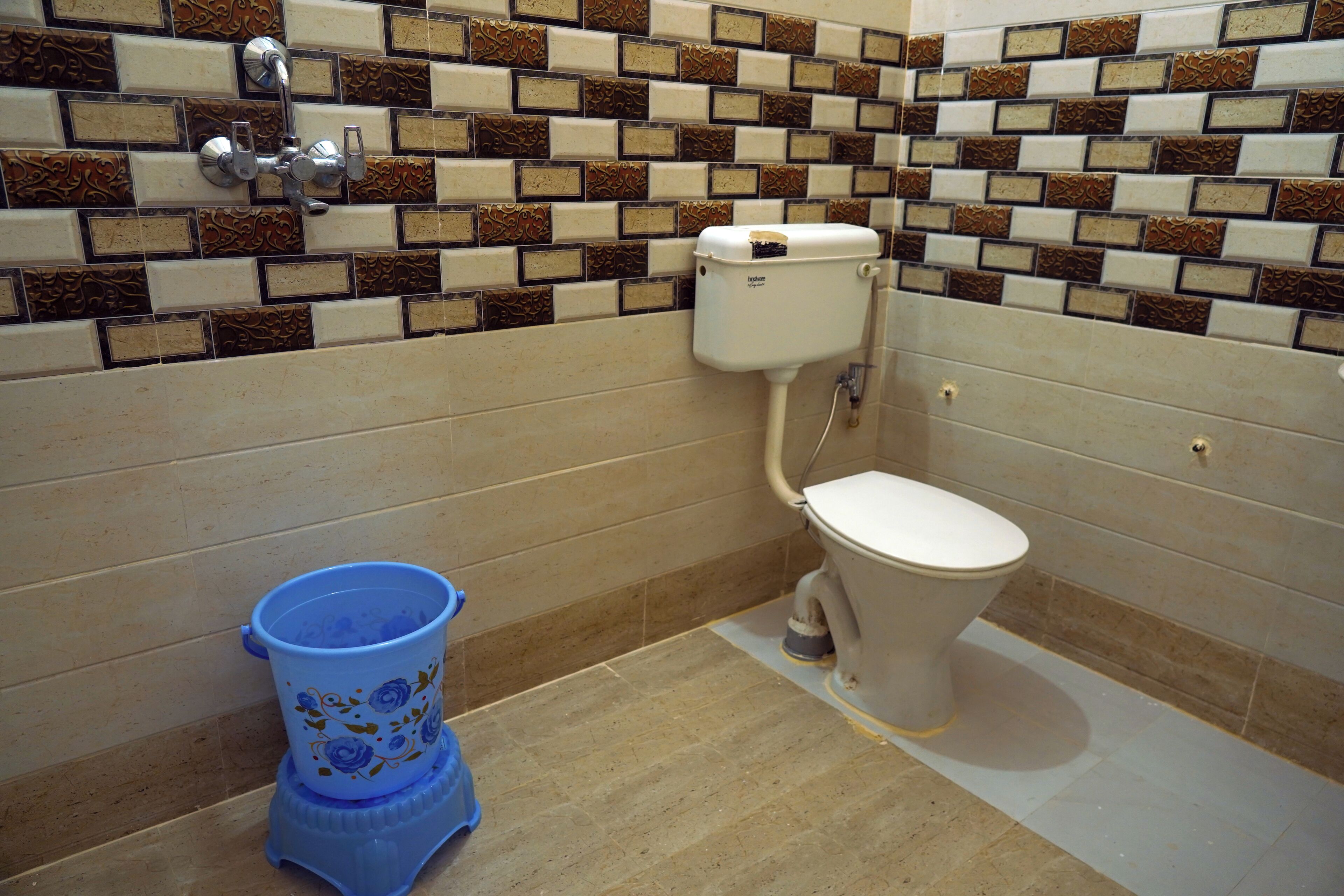 A bathroom with a toilet, a blue bucket, and a tiled wall with a brick pattern.