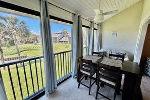 Ein Balkon mit Tisch und Stühlen, einem Deckenventilator und Blick auf eine angelegte Grünfläche mit Palmen und einem Gebäude im Hintergrund.