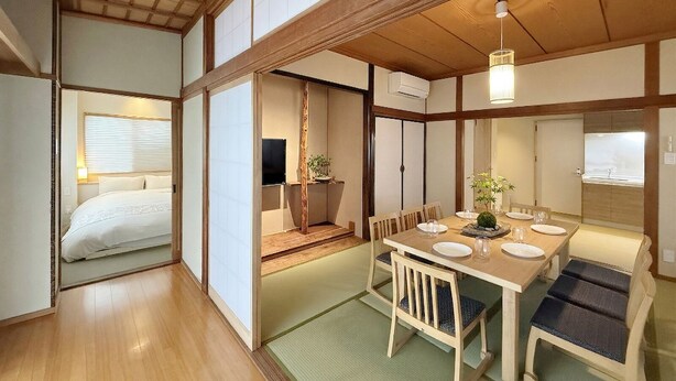 A traditional Japanese-style room with tatami flooring, a low table with chairs, a bed with white bedding, and sliding doors. - Namiki Hills (Hiroshima)