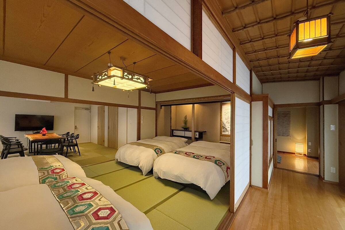 Una habitación tradicional japonesa con suelo de tatami, dos camas, una zona de comedor con mesa y sillas, y un televisor.