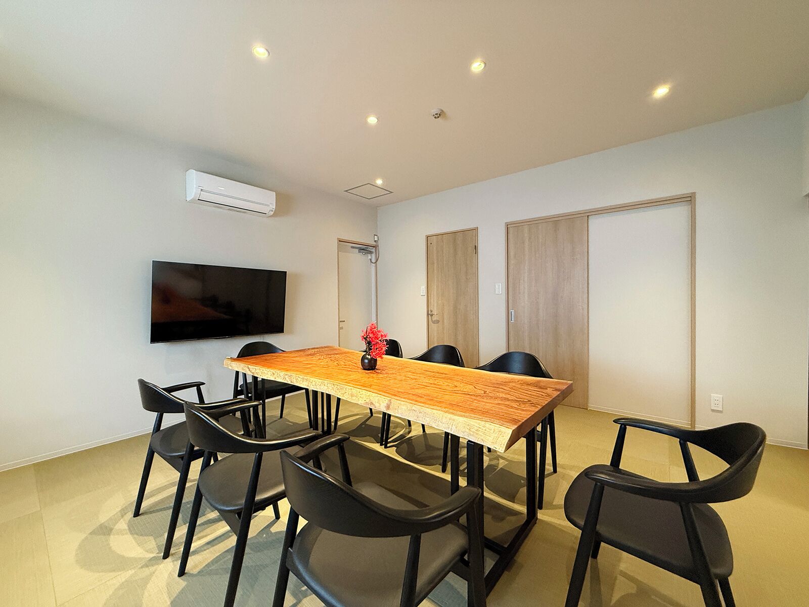 A conference room with a wooden table, black chairs, a flat-screen TV, and a ceiling-mounted air conditioner.